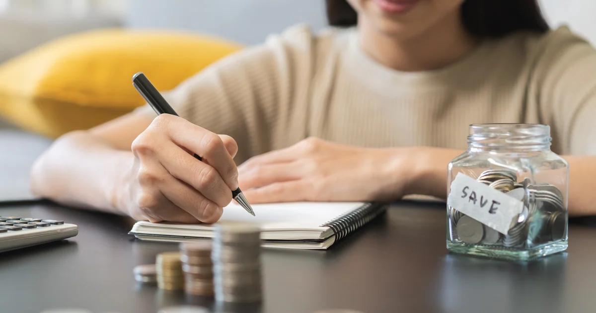 Mãos anotando em um caderno ao lado de um pote de poupança com a etiqueta SAVE